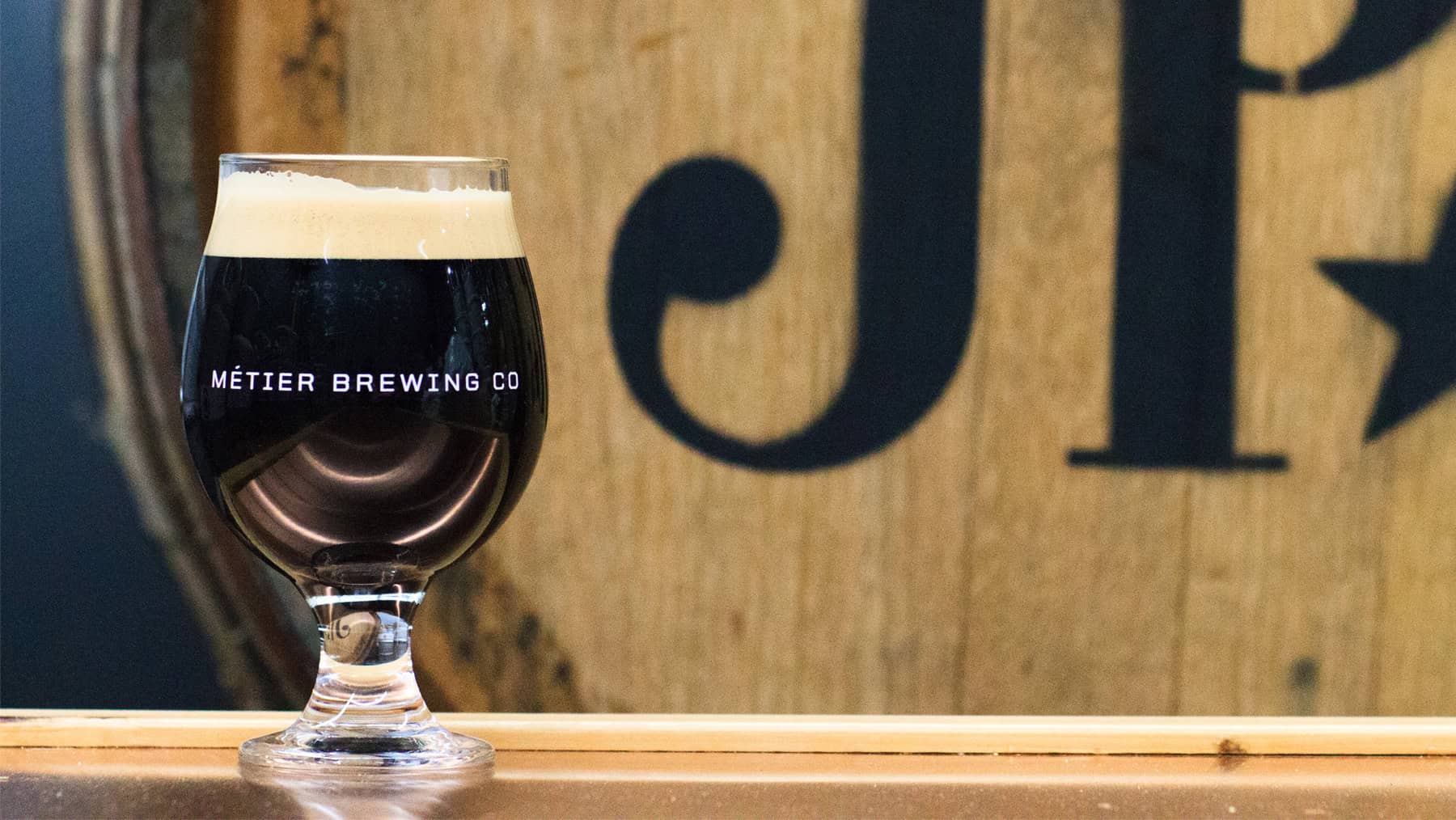 A short glass of dark beer on a counter in front of a barrel