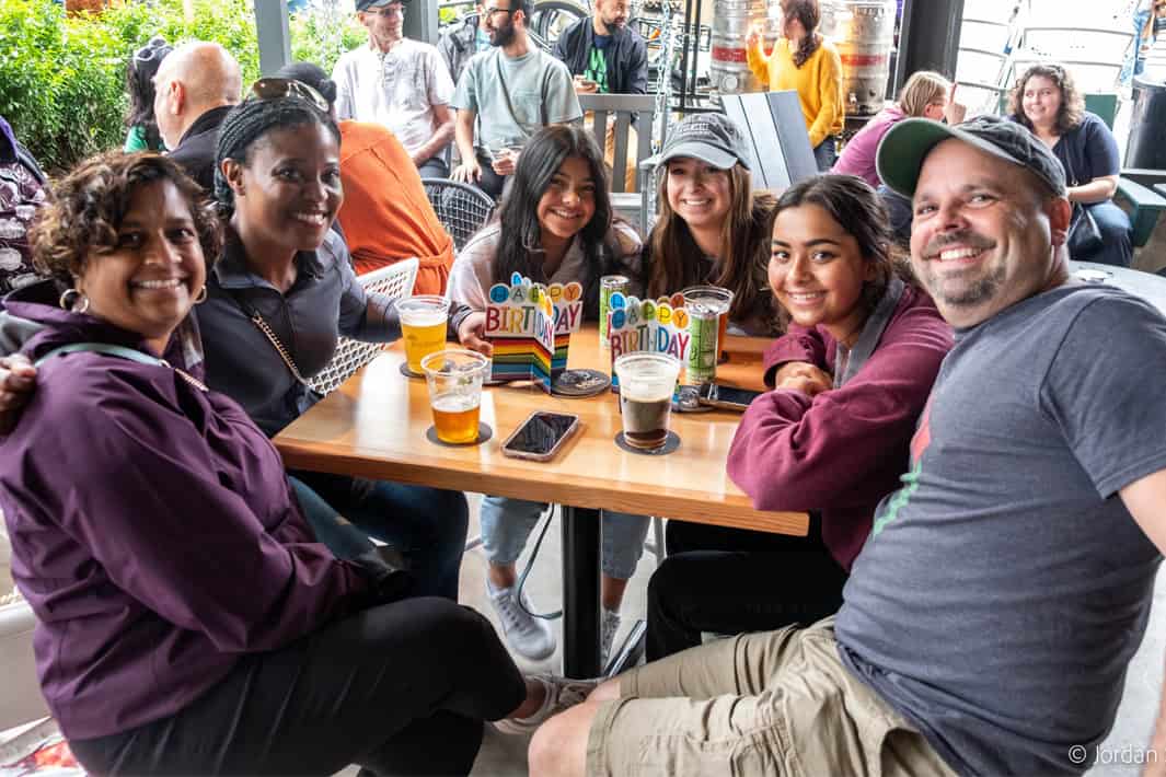 A group of six people smiling at the camera and sitting at a table with two 'Happy Birthday' cards and some beers.